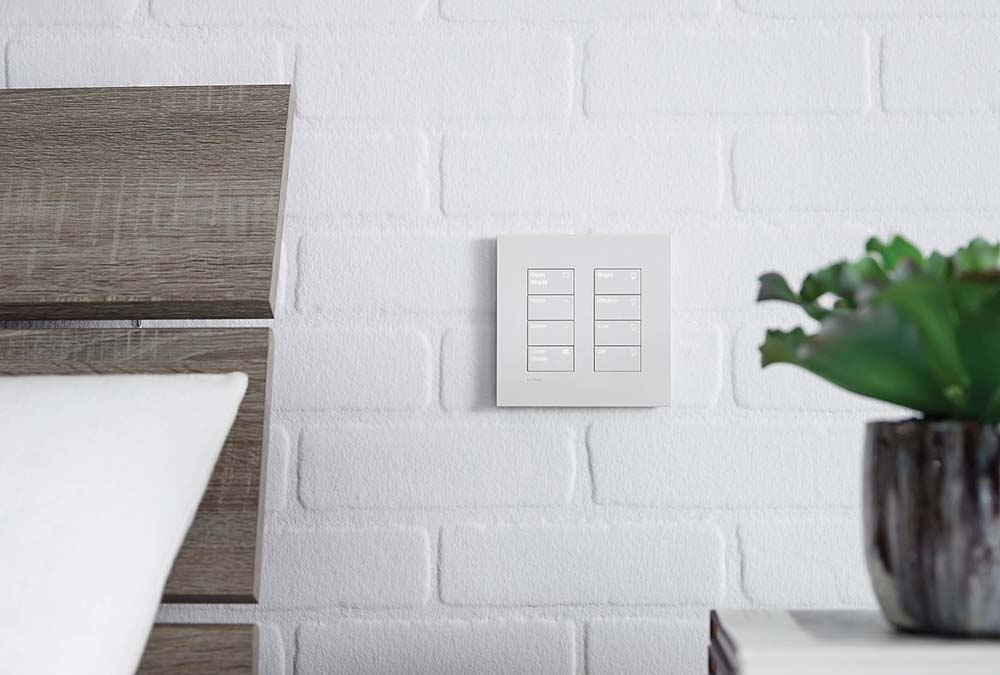 Modern bedroom with a white brick wall, wooden headboard, and a square smart control panel.