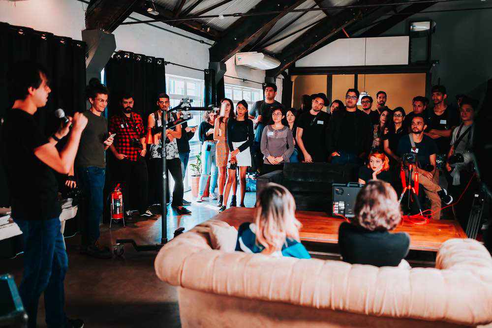 A speaker holding a microphone faces a diverse group of seated and standing attendees in a bright industrial loft space.