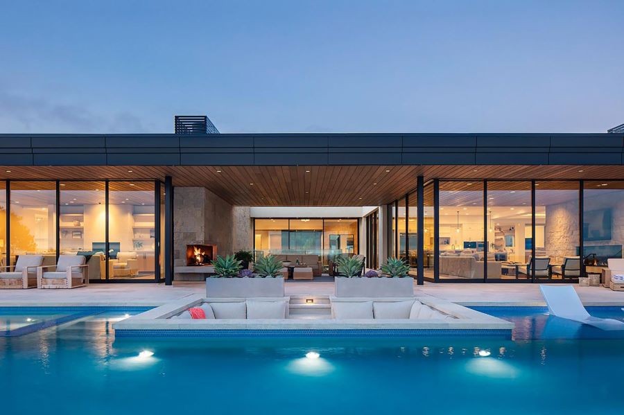 Looking over a pool into a home with floor-to-ceiling windows and beautiful lighting. 