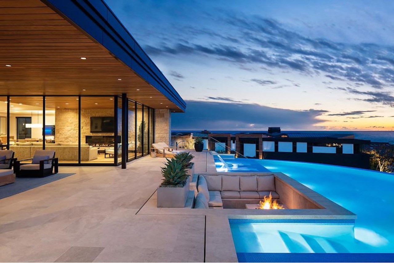 A backyard with a pool by the ocean. Glass walls reveal a softly lit living room and open-concept floor plan.