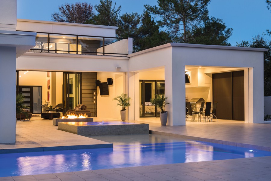 Modern white house in the evening with open windows and doors and a pool in the foreground.